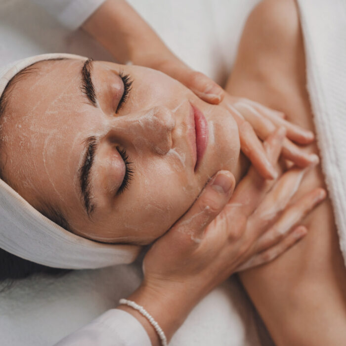 Beautician's hands applying facial cleansing foam on woman's face massaging skin in cosmetology clinic. Cosmetologist making skincare procedure in beauty salon, portrait of girl closeup.